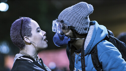Filmstill aus Espero tua revolta, zwei Menschen sehen sich in die Augen, eine Person trägt eine Gasmaske, nachts