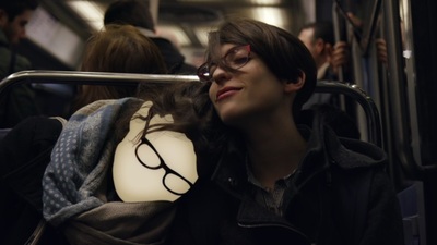 Zwei junge Frauen mit dunklen Haaren und Brillen sitzen nebeneinander in einer vollen U-Bahn auf einer Bank und lehnen sich zärtlich ineinander. Das Gesicht einer Frau ist hinter der schwarzen Brille wie eine weiße Wolke übermalt.
